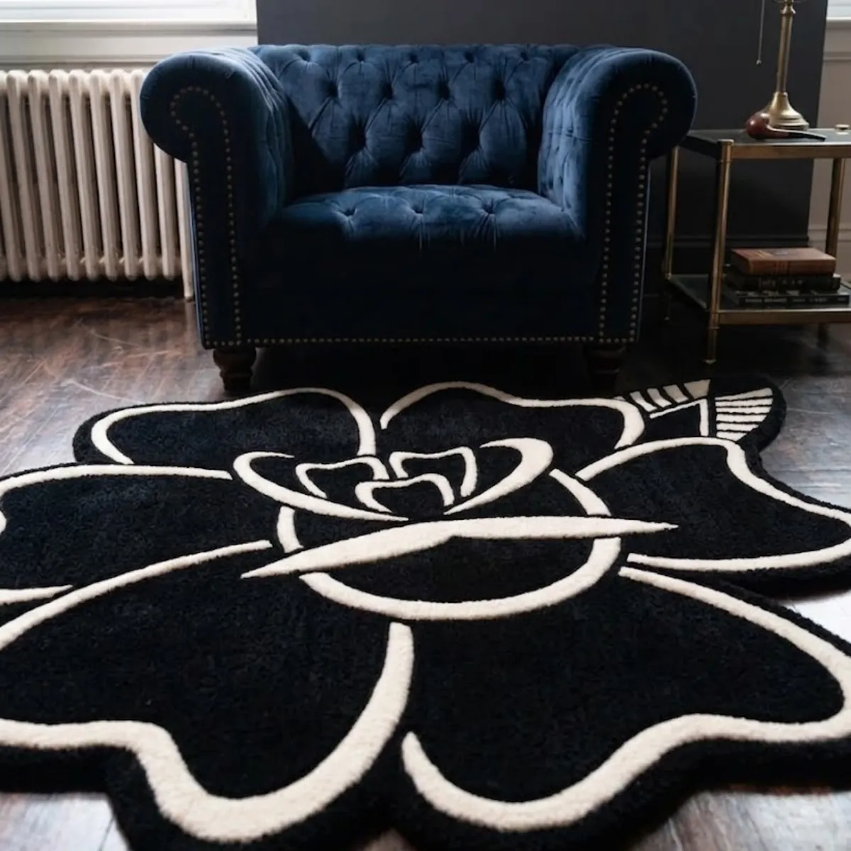 A low-angle view of the Blackwork Rose rug placed on a dark wood floor in front of a blue velvet armchair. This 8k quality shot highlights the bold, high-contrast white and black design of the traditional tattoo rose. Crafted with premium New Zealand wool and viscose, the thick tufted yarn features internal stuffing for a luxurious, natural softness, completely supported by a practical anti-slip backing.