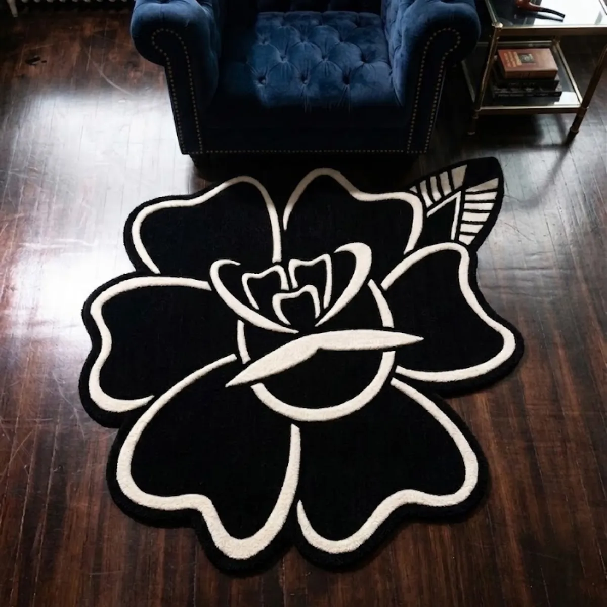 An overhead view of the Blackwork Rose rug resting on a dark hardwood floor. The custom cut-out hand-tufted design features a classic American Traditional tattoo aesthetic, showcasing a large flat black rose and a single leaf defined by thick, crisp white outlines. Designed as a flat 2D art piece with no color shading or gradients, the rug is styled in a room setting directly in front of a deep blue tufted velvet armchair.