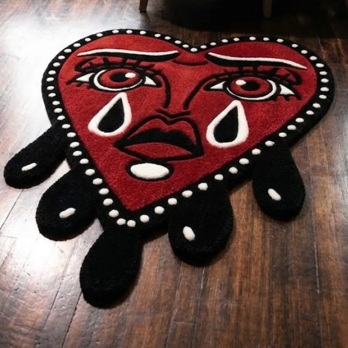 An angled close-up view of the Crying Heart rug resting on a dark wood floor. This 8k quality image highlights the crisp white and black accents of the face and eyes. The thick tufted yarn features internal stuffing for natural softness. This high-quality construction perfectly captures the bold color blocking of the custom-shaped American Traditional piece.
