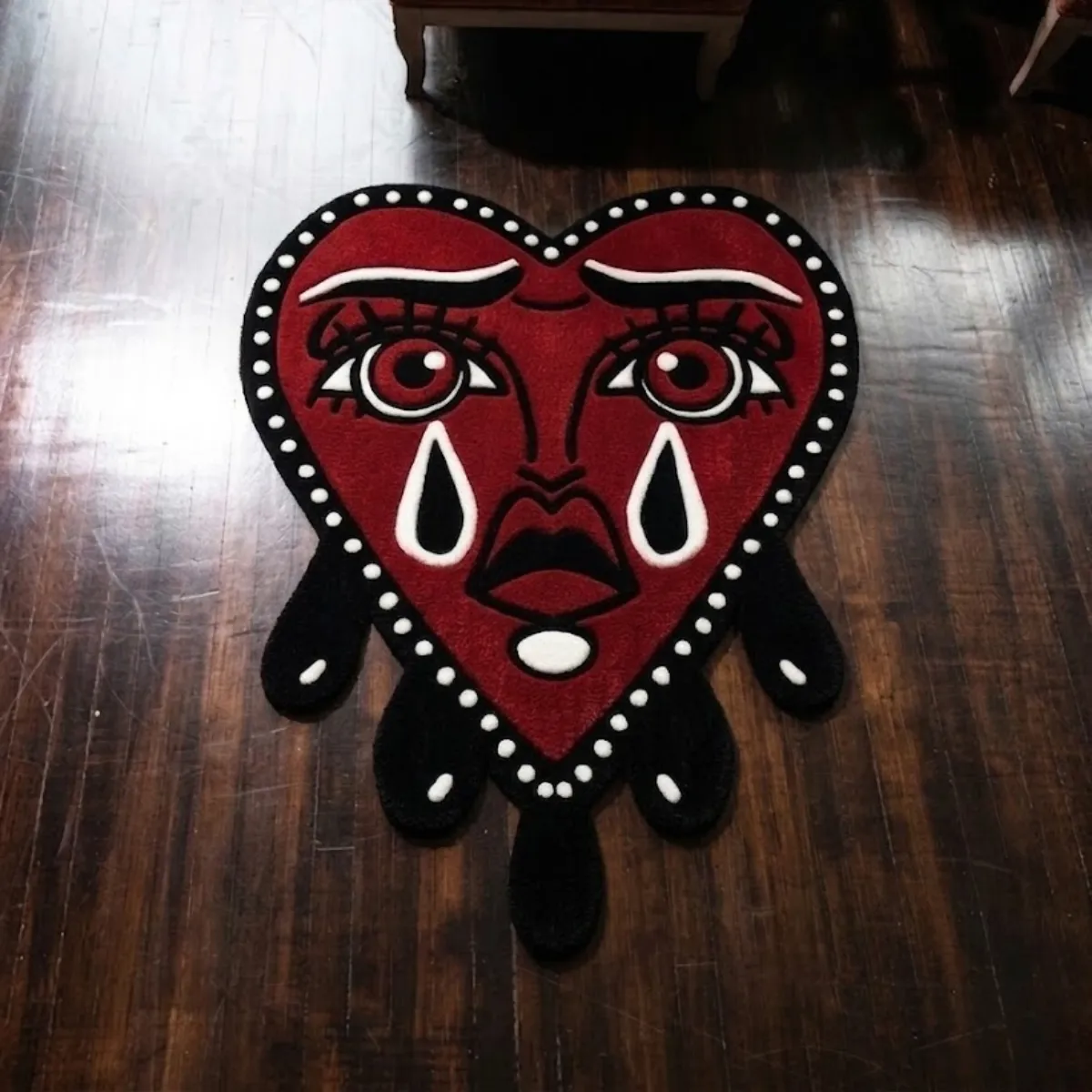 An overhead view of the red Crying Heart rug resting on a dark hardwood floor. The custom cut-out design features an American Traditional tattoo aesthetic. It showcases a large face with bold black outlines and black teardrops falling from the bottom edge. Designed without color shading or gradients, the rug is crafted from New Zealand wool and viscose and includes an anti-slip backing.