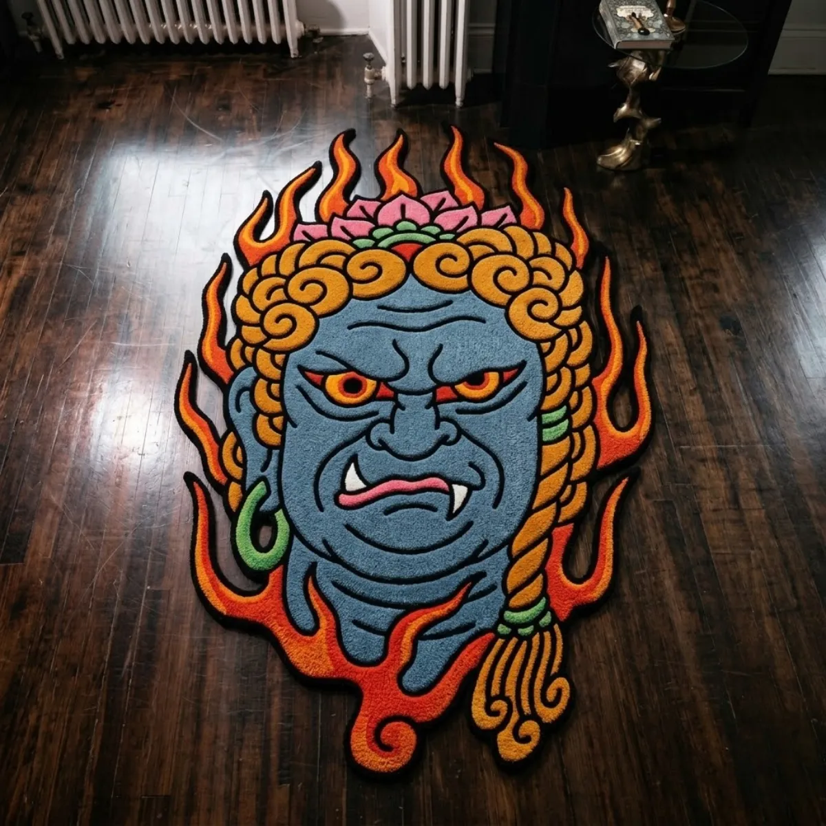 An overhead view of the Fudo Myoo rug resting on a dark hardwood floor. The custom cut-out hand-tufted design features a bold Japanese Traditional tattoo aesthetic, showcasing a fierce blue deity face with a pink lotus crown, green earrings, and vibrant orange and red flames. Designed as a flat 2D art piece with no color shading or gradients, the statement rug is expertly crafted from New Zealand wool and viscose and includes a practical anti-slip backing.