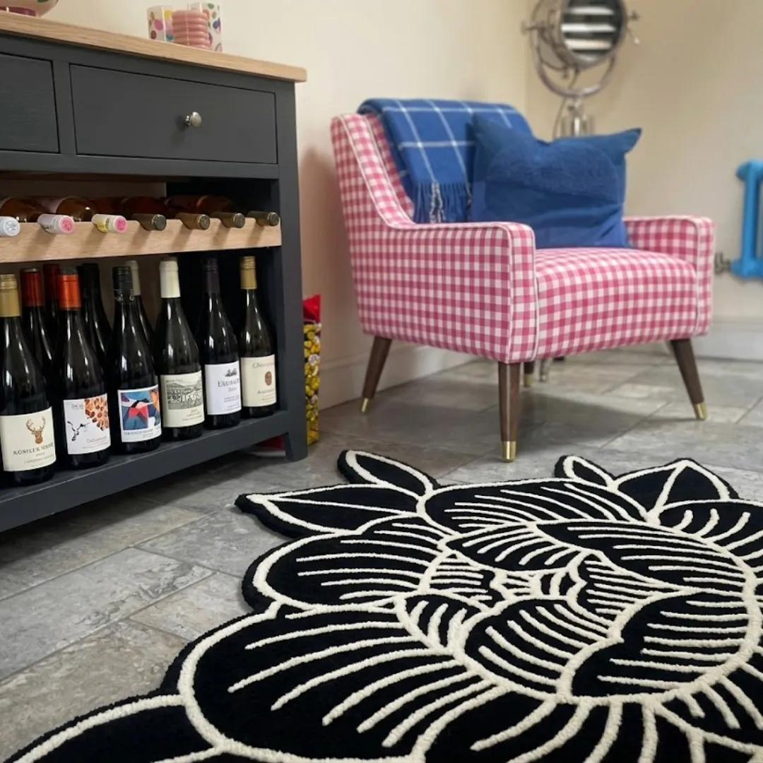 A low-angle close-up of the black and white Peony Flower rug resting on a tiled floor next to a dark grey wine cabinet. The detailed shot highlights the thick, plush tufted yarn texture of the traditional Japanese floral design, with a pink checkered chair softly blurred in the background.