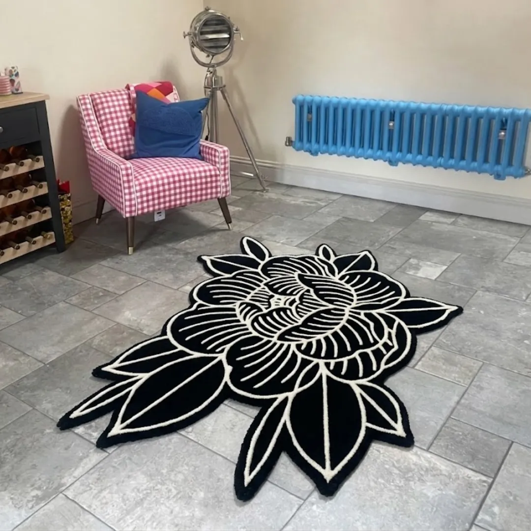 An angled view of the hand-tufted Peony Flower rug placed on a grey tiled floor. The striking black rug features bold white line work outlining a classic Japanese tattoo-style peony blossom, styled in a room next to a pink checkered armchair, a tripod lamp, and a bright blue radiator.