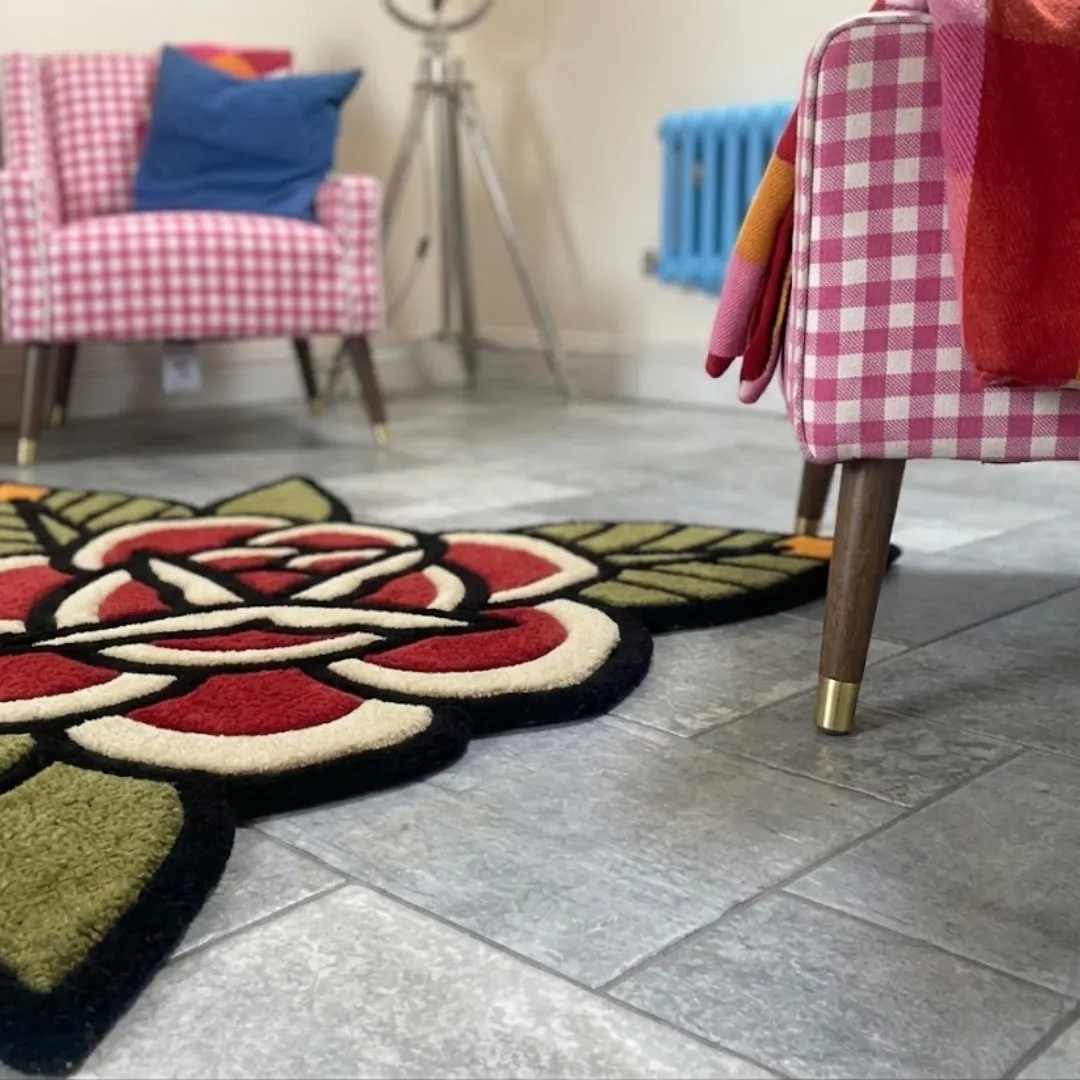 A low-angle close-up of the Red Rose Flower Rug resting on a grey tiled floor. The detailed shot highlights the thick, plush tufted yarn texture of the red, cream, and green floral design, emphasizing the crisp black outlines typical of American traditional tattoo art, with the legs of a pink checkered chair in the foreground.