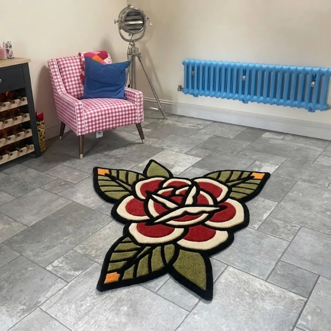 An angled view of the Red Rose Flower Rug placed on a grey tiled floor. The hand-tufted design features a classic American traditional tattoo style with a bold red blossom, cream-colored inner petals, and green leaves outlined in black, styled in a room next to a pink checkered armchair and a bright blue radiator.