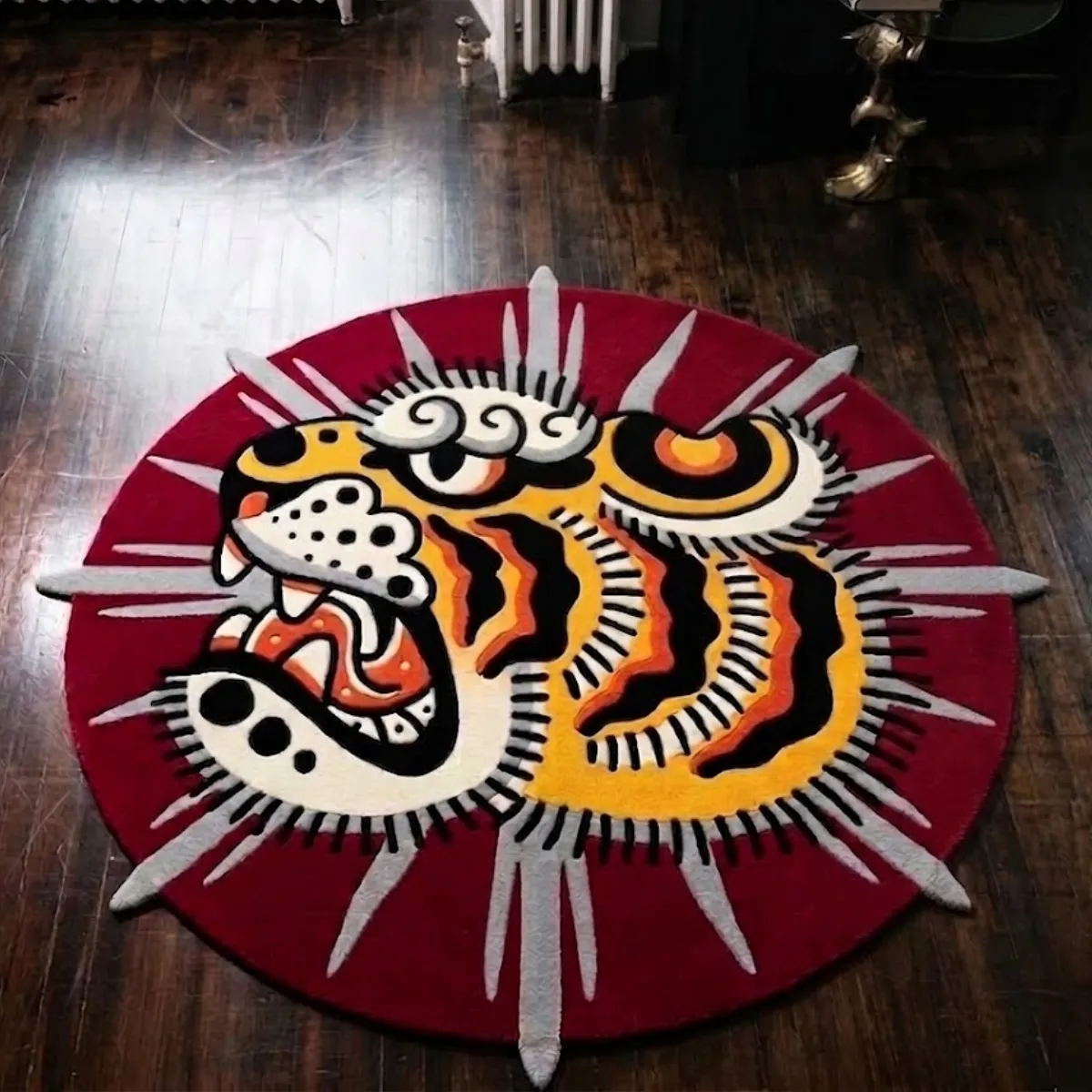 An overhead view of the Sun Tiger rug resting on a dark hardwood floor. The custom hand-tufted design features a bold traditional tattoo aesthetic, showcasing a yellow, black, and orange tiger head in profile against a solid red circular background, with grey sun rays extending playfully past the edges. Designed as a flat 2D art piece with no color shading or gradients, the rug is expertly crafted from New Zealand wool and viscose and includes a practical anti-slip backing.