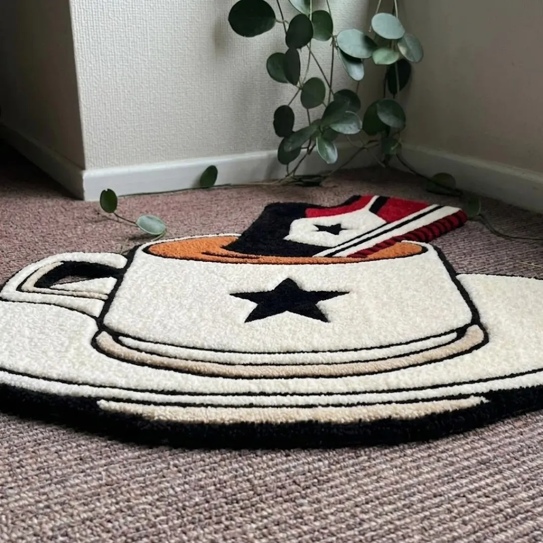 An angled perspective shot of a hand-tufted sneaker teacup rug, highlighting its playful novelty silhouette and plush tufted wool texture on a carpeted floor.