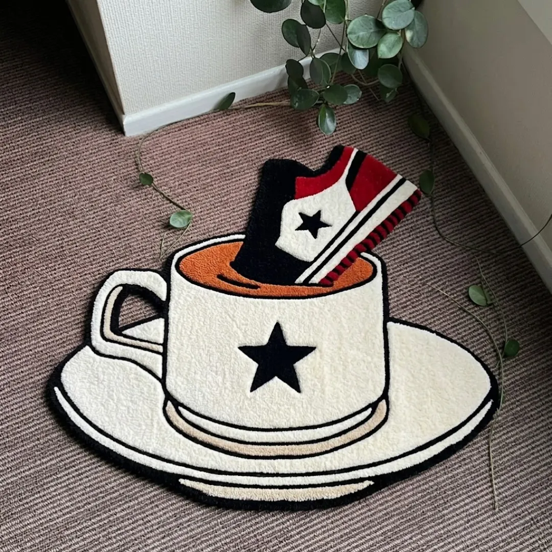 A top-down studio product photograph of a unique novelty area rug shaped like a teacup and saucer with a red, black, and white sneaker dunked inside, displayed on a carpeted floor.