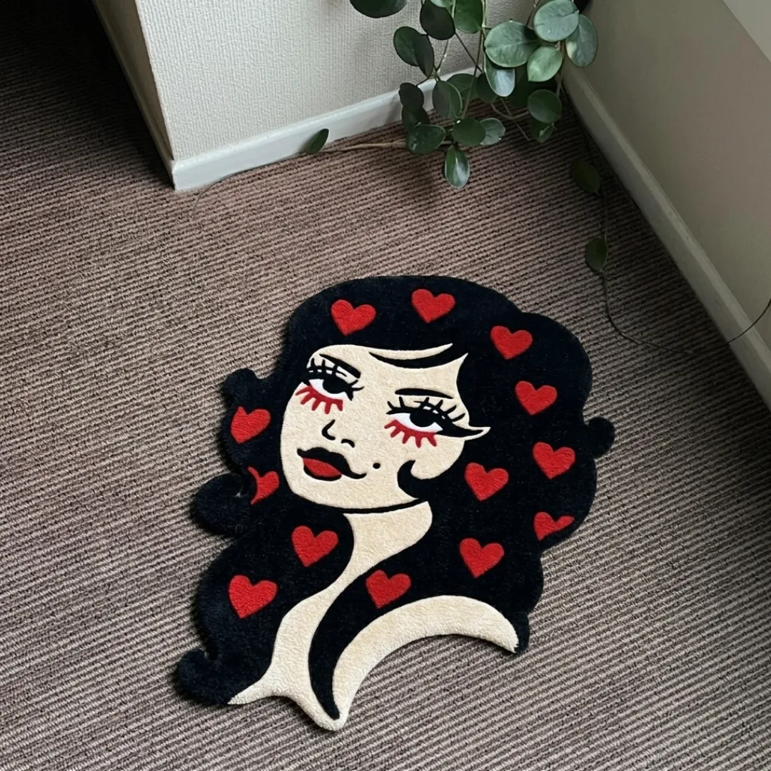 A top-down studio product photograph of a unique, woman-shaped area rug featuring a black-haired lady with red lipstick and multiple red hearts scattered in her hair, displayed on a textured carpet.