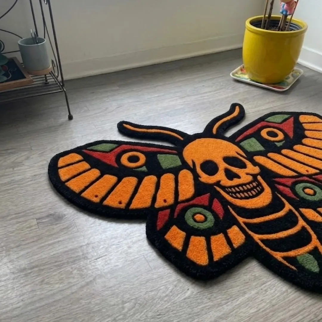 An angled perspective shot of a hand-tufted orange death head moth rug, highlighting its expressive moth silhouette and the high-quality wool texture on a wooden floor.