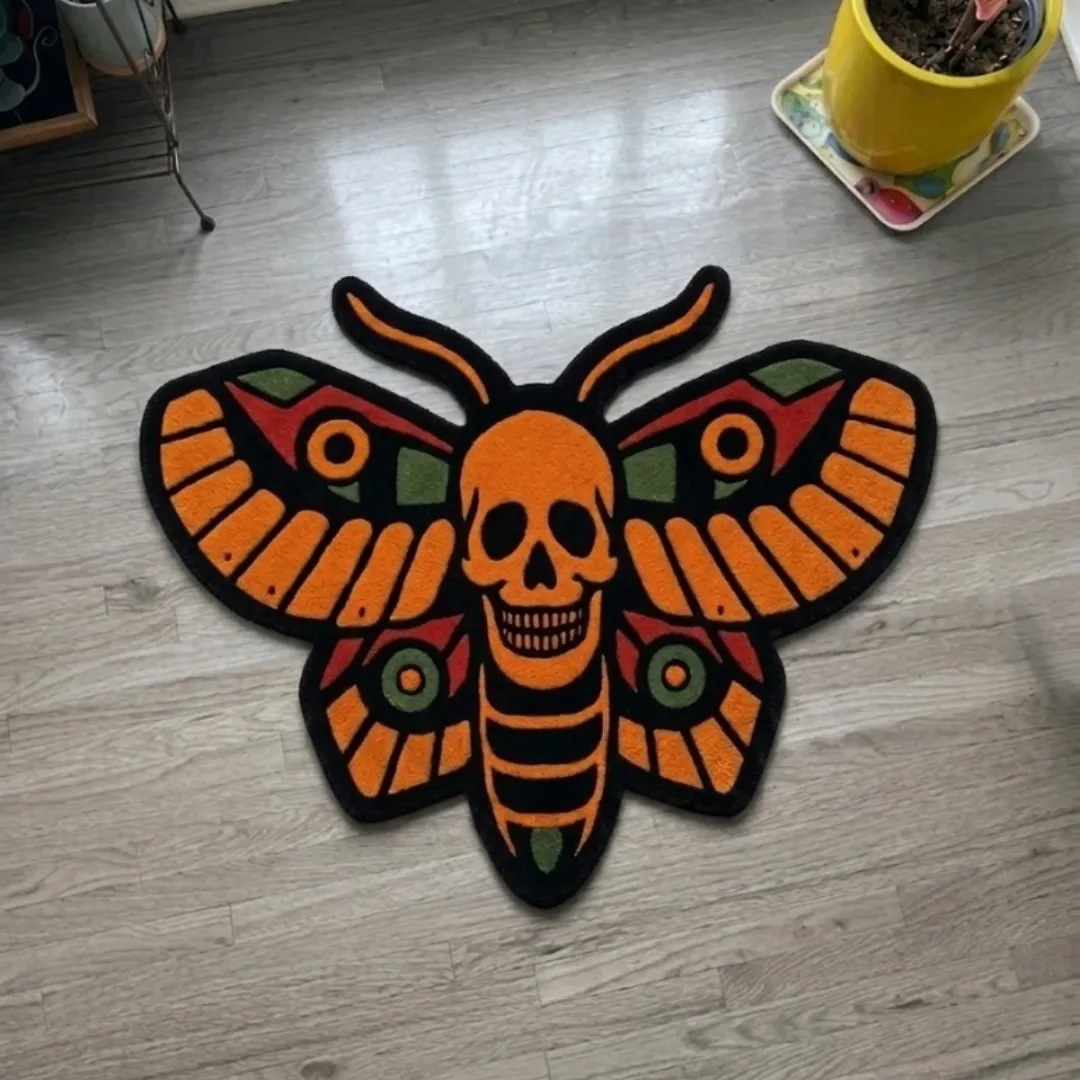 A top-down studio product photograph of a unique, moth-shaped area rug featuring a central skull motif and detailed orange, red, and green wing patterns, displayed on a wooden floor.