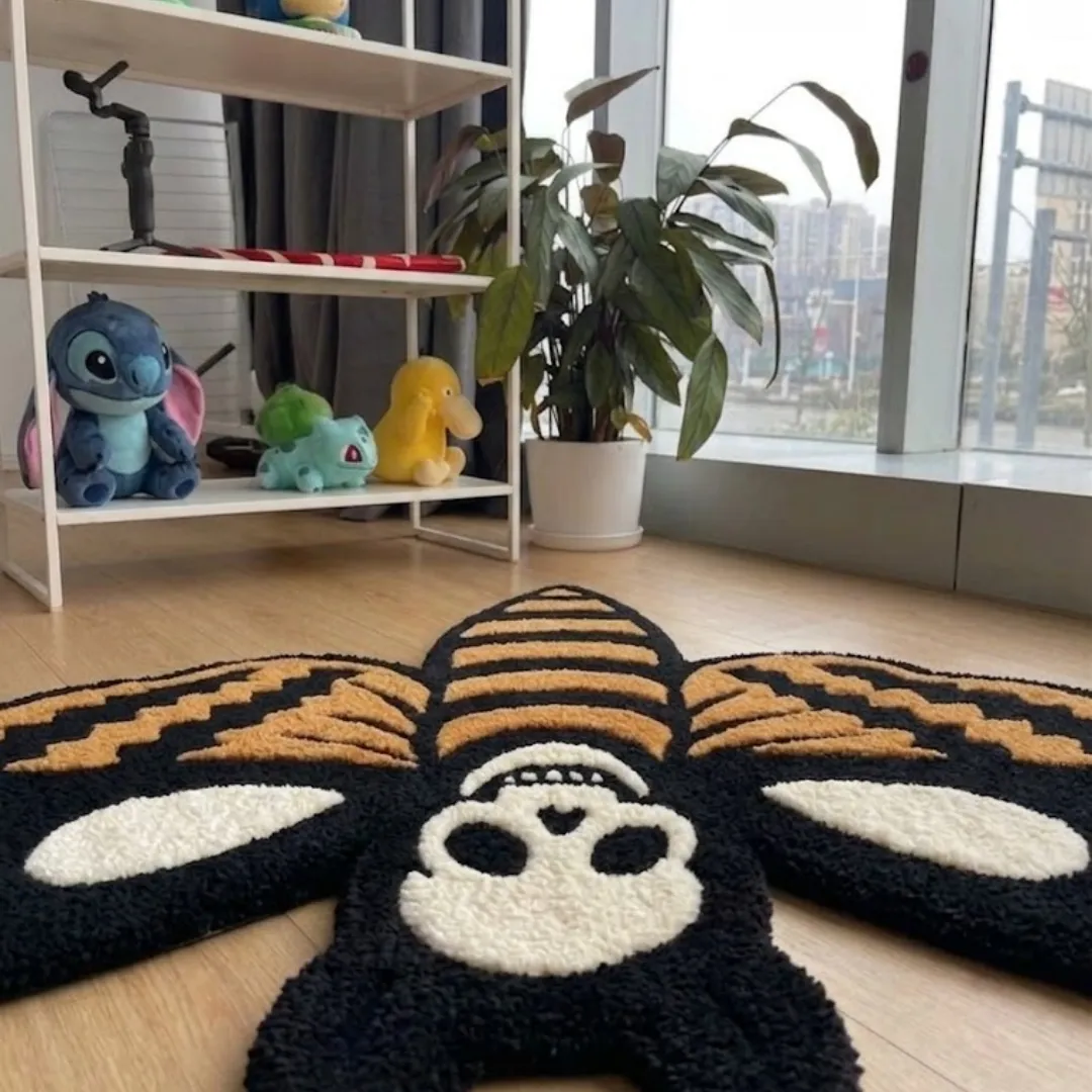 An interior lifestyle shot of a moth-shaped rug with a skull design, styled on wooden flooring in a room near a shelf with character collectibles.