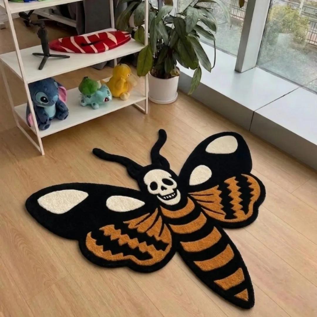 A top-down studio product photograph of a unique, moth-shaped area rug featuring a central skull motif and detailed orange, black, and cream wing patterns, displayed on a wooden floor.