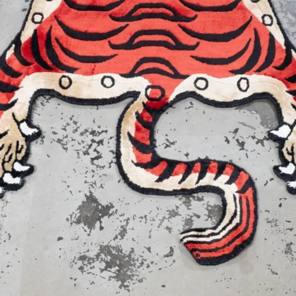 Red Tibetan-style tiger rug with bold black stripes, blue eyes, and cream edges placed on a textured concrete floor.