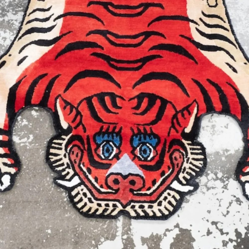 Red Tibetan-style tiger rug with bold black stripes, blue eyes, and cream edges placed on a textured concrete floor.