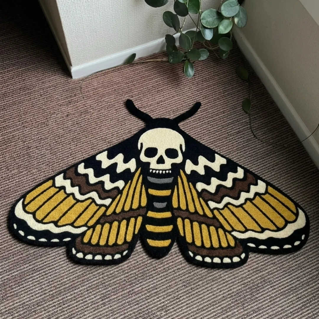 A top-down studio product photograph of a unique, moth-shaped area rug featuring a central skull motif and detailed golden-yellow wing patterns, displayed on a textured carpet floor.