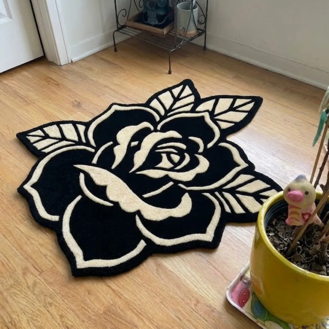 An interior lifestyle shot of a black and cream rose-shaped area rug placed on wooden flooring, accented by a yellow potted plant and a metal storage shelf.