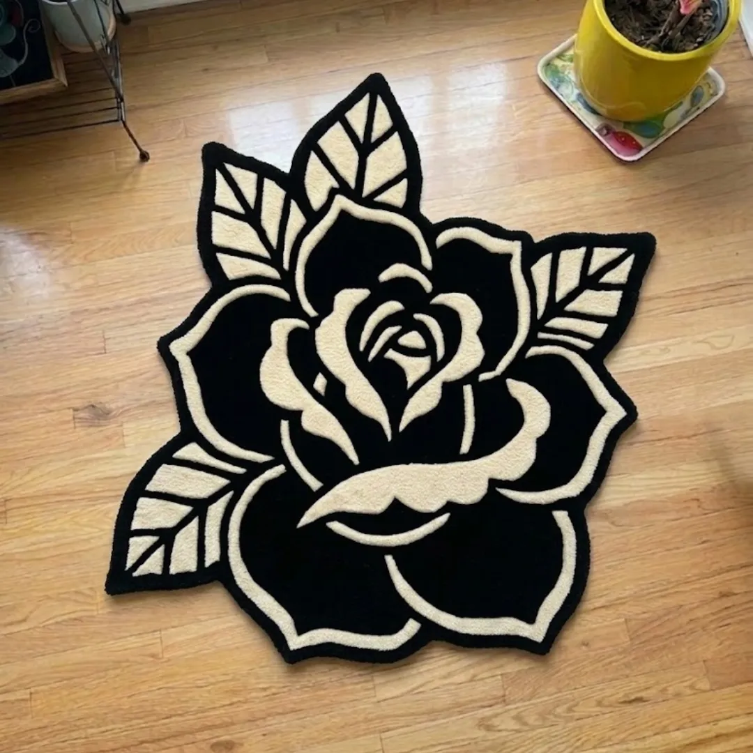 A top-down studio product photograph of a black and cream rose-shaped area rug featuring bold, graphic lines inspired by traditional tattoo art, displayed on a wooden floor.