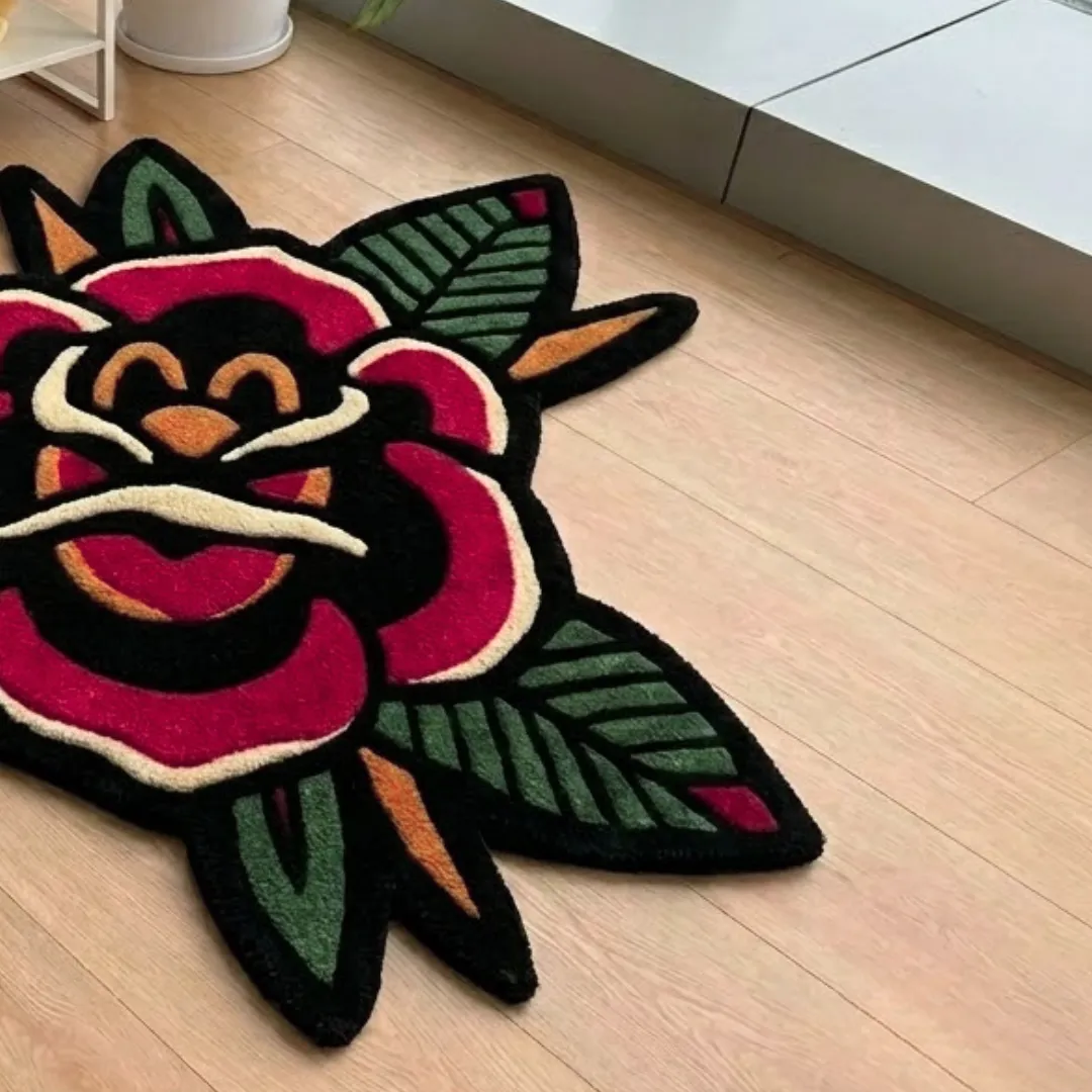 A perspective view of a bold rose-shaped area rug with vivid red petals, green leaves, and gold accents, displayed on light wooden flooring.