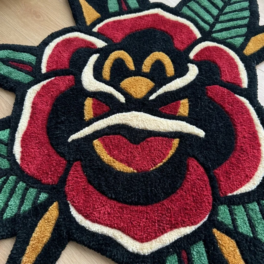 A detailed close-up of a hand-tufted rose area rug featuring bold black outlines, rich red petals, and green leaf details in a traditional tattoo art style.