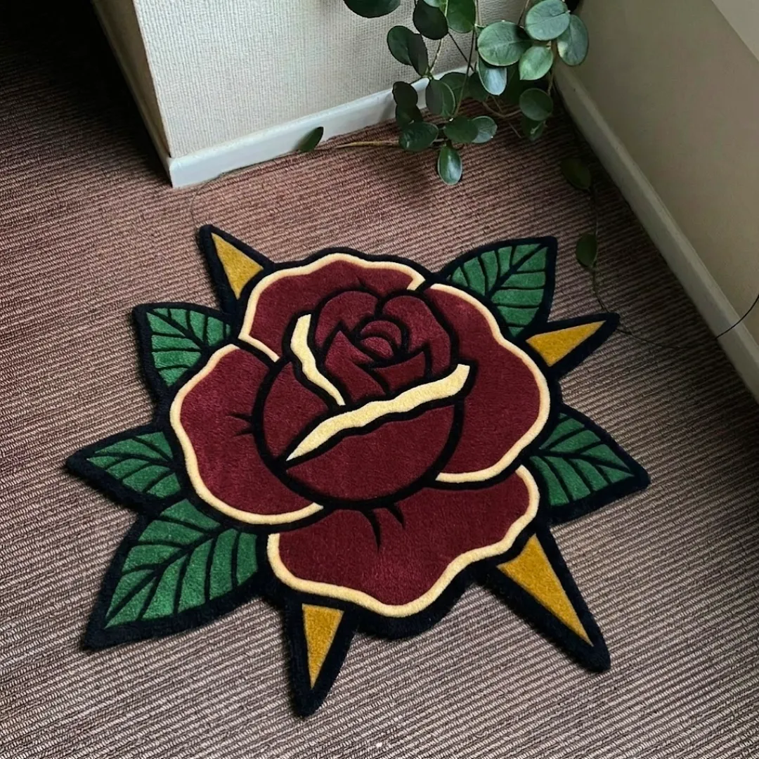 A top-down product photograph of a uniquely shaped area rug featuring a traditional tattoo-inspired red rose design with green leaves and yellow accents, placed on a textured carpet.