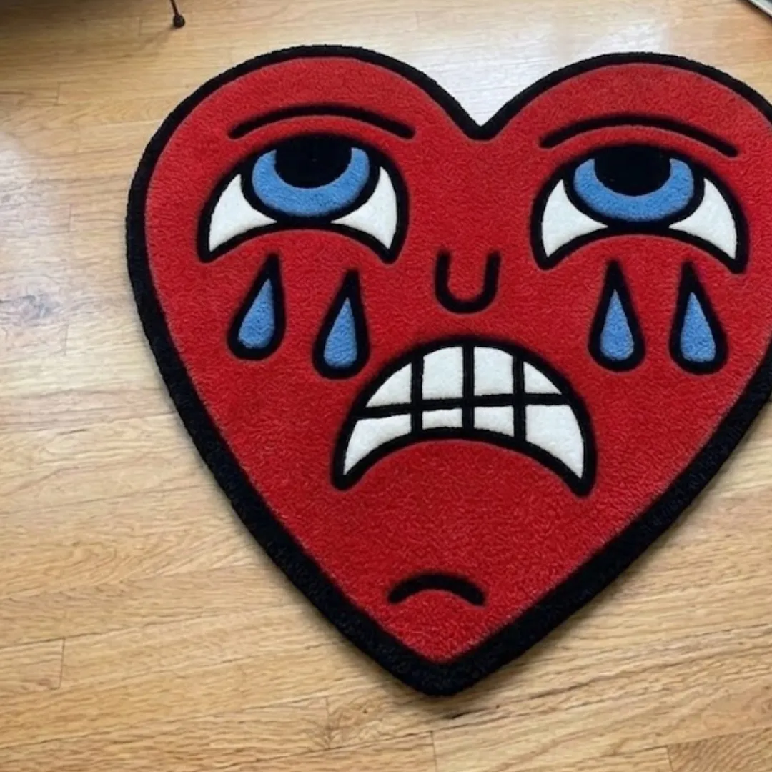 An interior lifestyle shot of a red heart-shaped area rug with a crying face design, styled on light hardwood flooring in a colorful home setting.