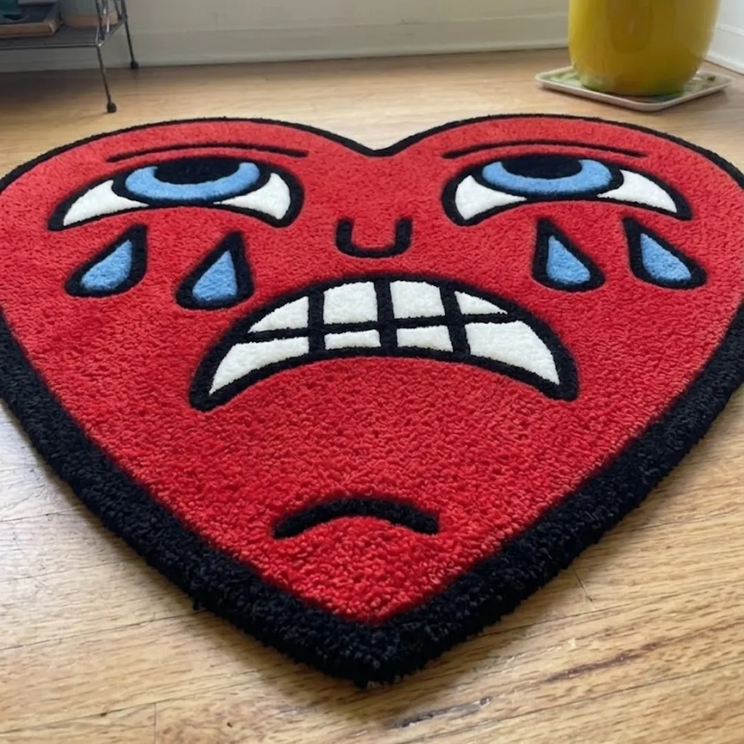 An angled perspective shot of a hand-tufted crying heart rug, highlighting its expressive cartoon-style face and the dense, high-quality tufted wool texture on a wooden floor.