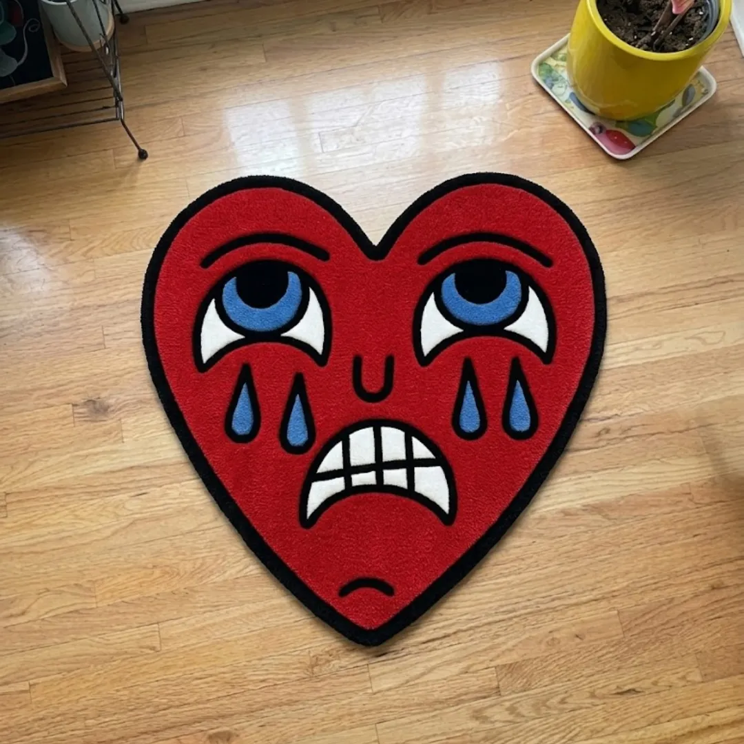 A top-down studio product photograph of a unique, heart-shaped area rug featuring a playful, expressive "crying" character face in red, blue, and white, displayed on hardwood flooring.