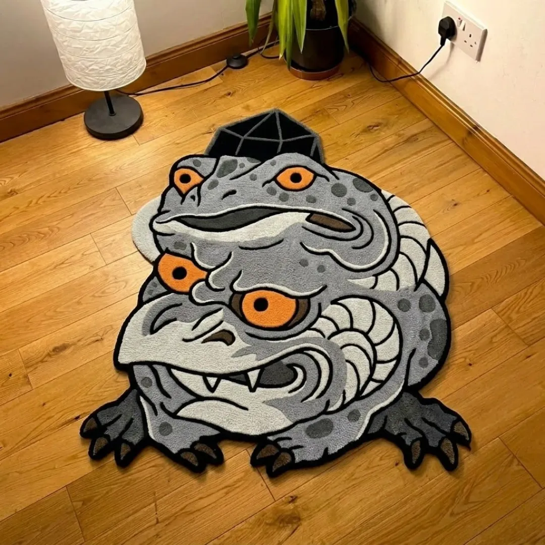 A top-down studio product photograph of a unique, quirky frog demon-shaped area rug featuring a two-headed creature design in shades of grey with orange eyes, displayed on hardwood flooring.