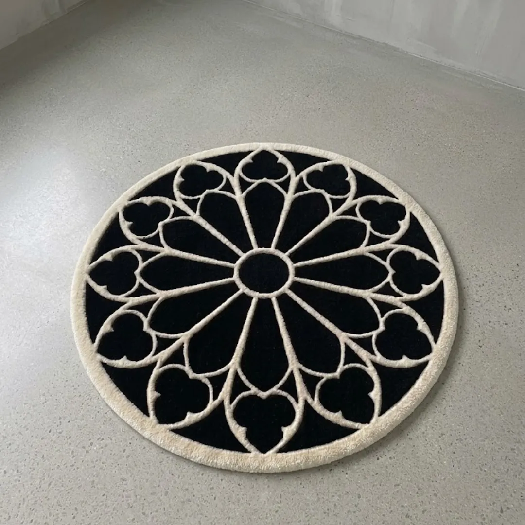 A full top-down view of the round Eclipse Halo Rug resting on a light grey concrete floor. The hand-tufted rug features a striking black background with a contrasting white geometric gothic rose window pattern and a thick white outer border.