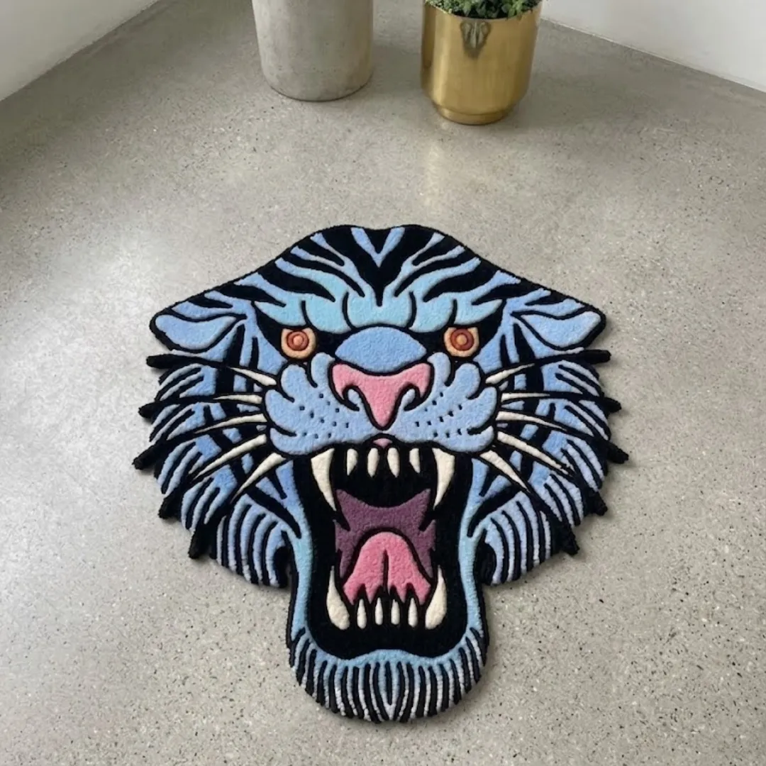 A top-down view of the Rebellion Blue Rug resting on a light speckled concrete floor. The hand-tufted rug features an American traditional tattoo style tiger head in light blue and black stripes, with a fierce open mouth, pink nose, and sharp white teeth.