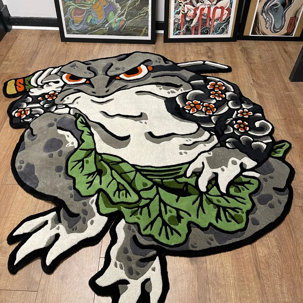 kaeru no bujin anime style hand tufted rug Eye level view of the Kaeru No Bujin rug in an art studio setting, showcasing the custom die-cut shape and bold black outlines of the samurai frog character against a backdrop of framed tattoo art.