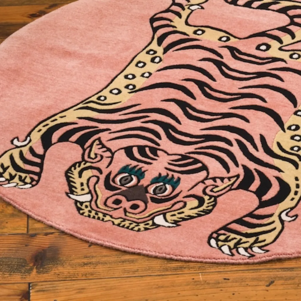 High angle view of the round Petal Prawl rug placed on a rustic wooden floor, featuring a traditional Tibetan tiger design adapted into a circular pink format with yellow accents.