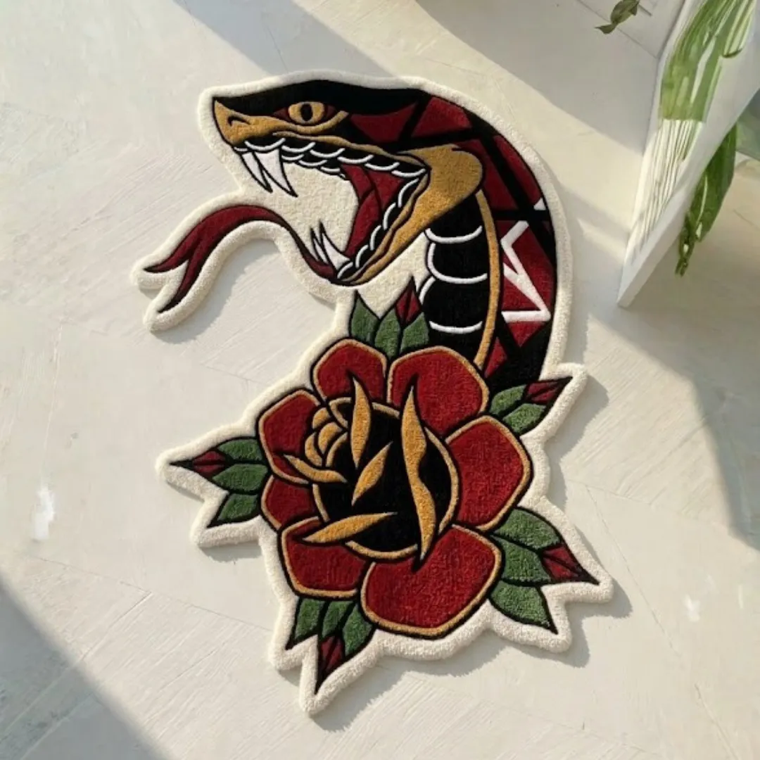 An angled view of the Snake Rose Traditional Rug resting on a bright white wooden floor bathed in natural sunlight. The hand-tufted rug features a classic American traditional tattoo design, showcasing a fierce open-mouthed snake with black, red, and white scales and a red forked tongue, intertwined with a bold red and gold rose with dark green leaves.