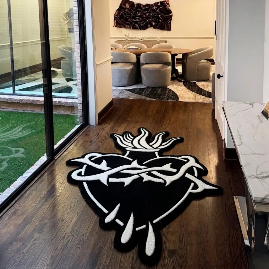 The black and white Thorned Heart Rug styled in a modern home hallway with dark wood floors. The custom cut-out tufted rug, featuring a striking sacred heart, thorns, and flames, is positioned near large glass sliding doors, leading the eye toward a bright, contemporary dining room.