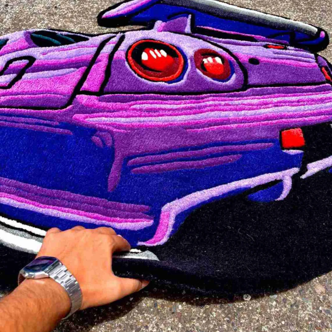 Perspective view of the Arrow Crest rug with a hand lifting the edge, showcasing the thick hand-tufted wool pile and the vibrant shading of the purple bumper.