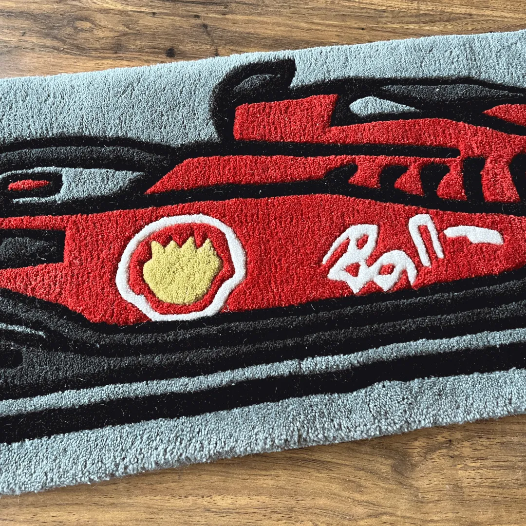 Close-up detail of the Asphalt Aura rug featuring the nose of a red Formula 1 style race car with stylized white and yellow logos on a grey background.
