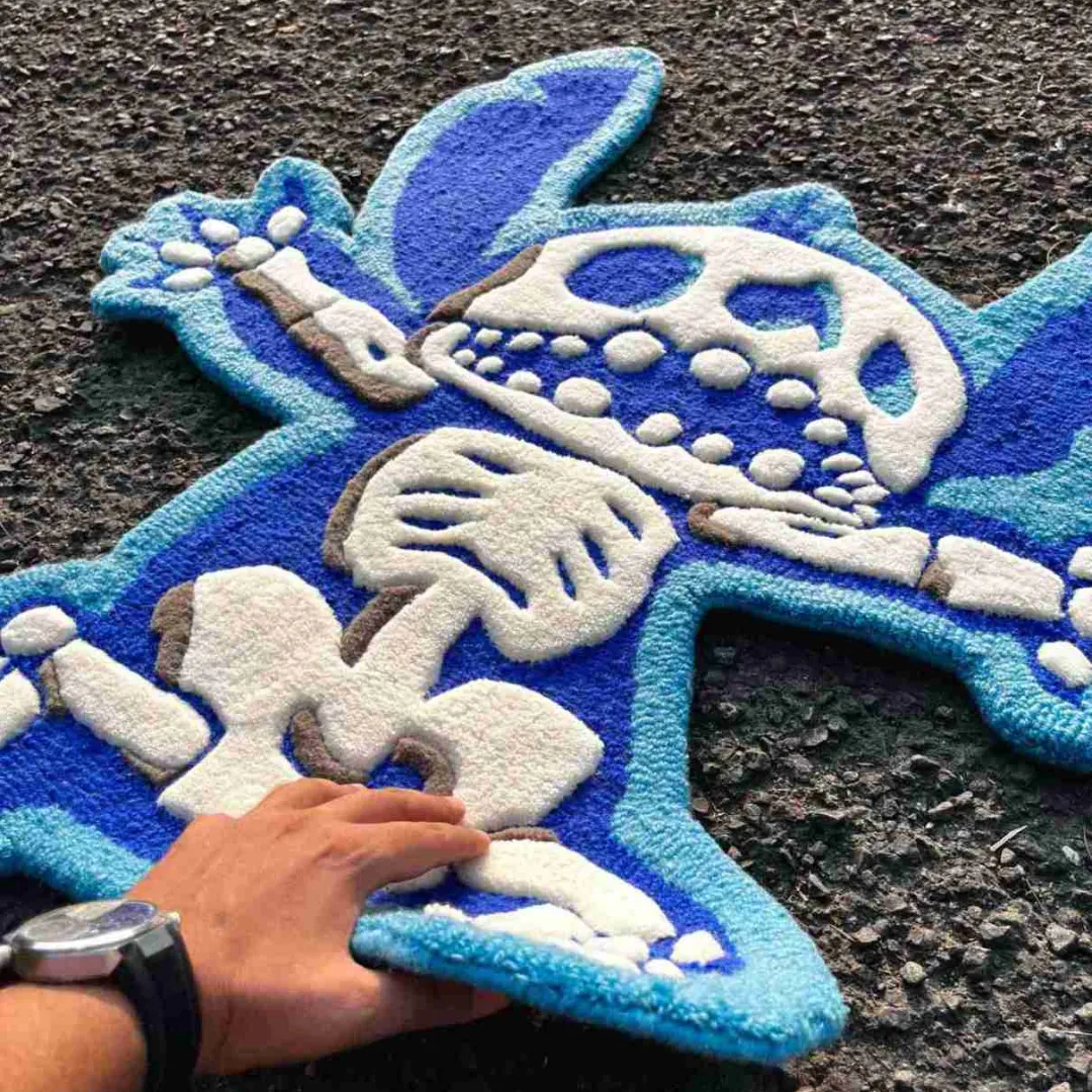 Angled view showing a hand lifting the bottom edge of the Azure Flux rug on an asphalt street, emphasizing the thick, flexible wool pile and the bold white skeleton design against the vibrant blue base.