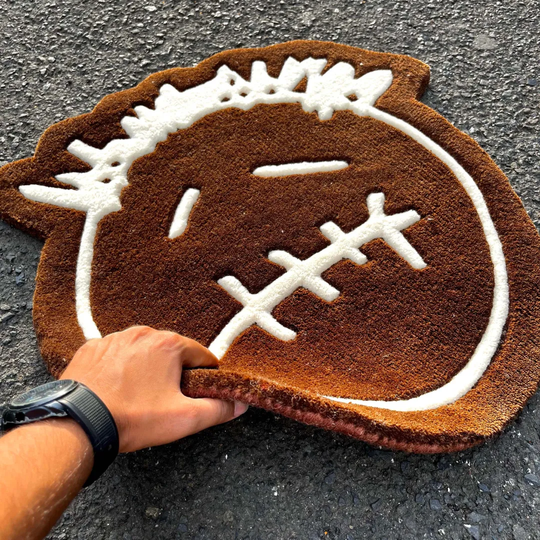 Angled view showing a hand lifting the bottom edge of the Bruma Mark rug on a concrete road, emphasizing the thick, flexible wool pile and the bold white line art of the streetwear-inspired face design.