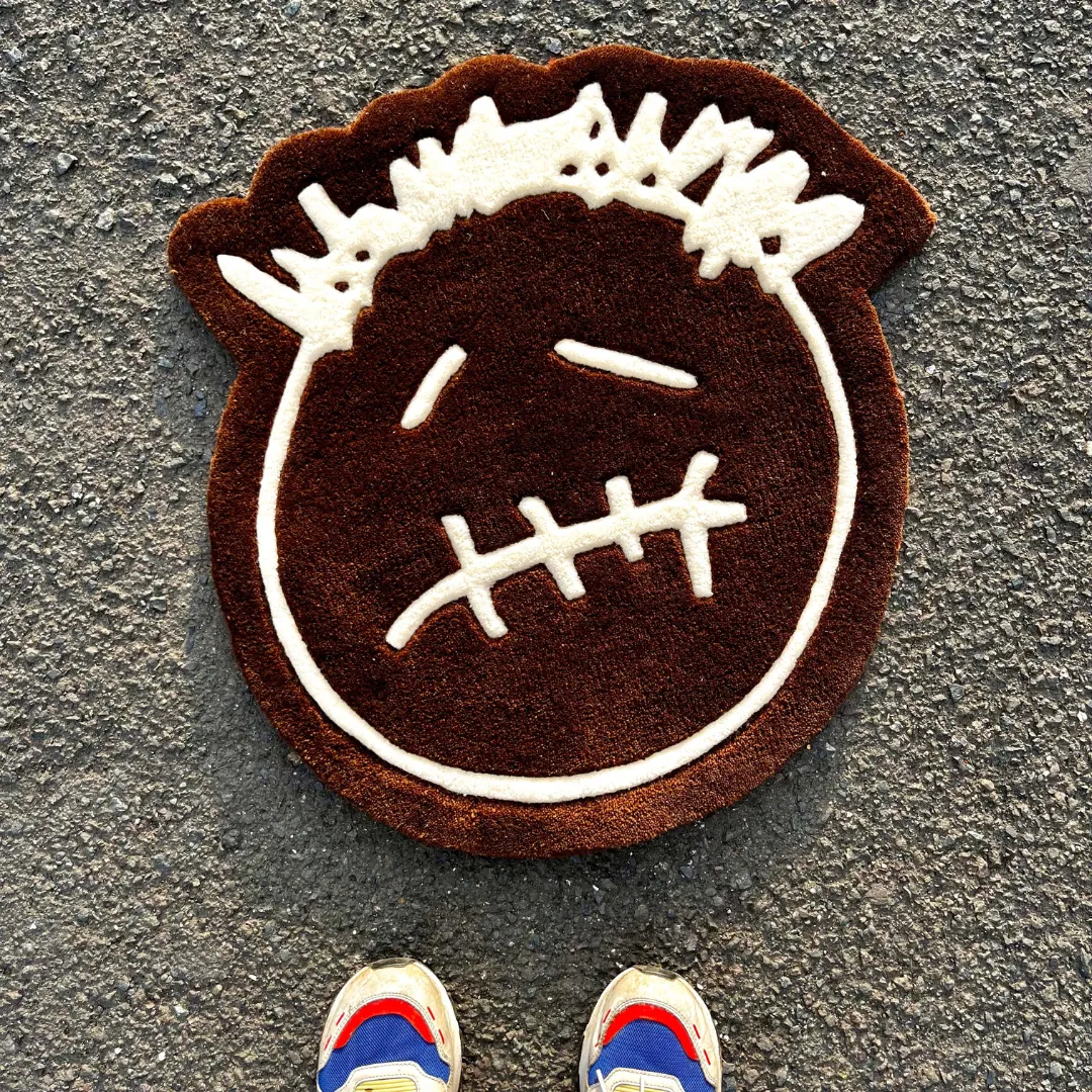 First-person POV lifestyle view of the Bruma Mark rug placed on a concrete street setting, showcasing the brown and white stylized face design alongside a pair of colorful sneakers at the bottom edge.