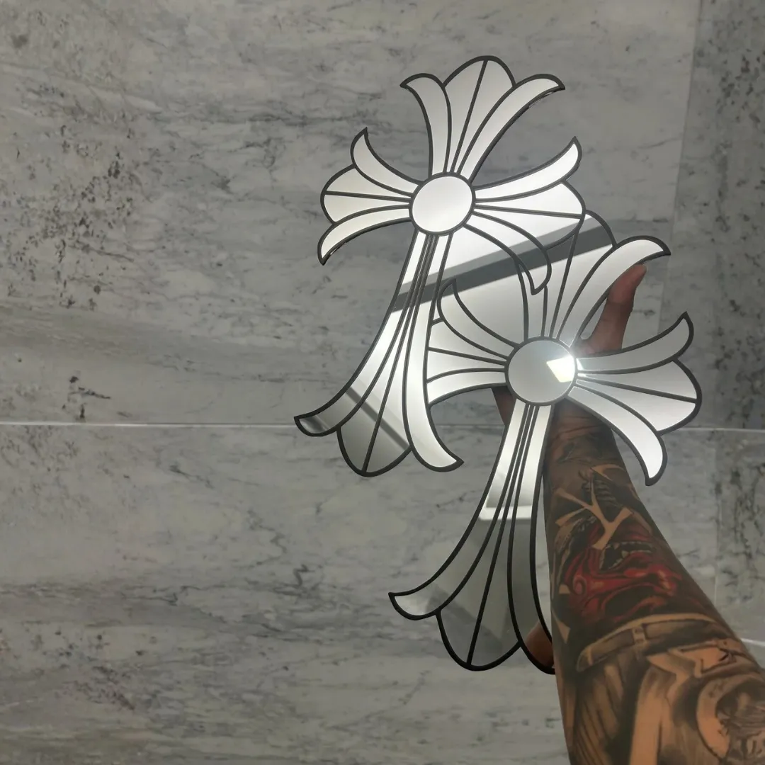 A tattooed hand holding up two gothic cross-shaped mirrors against a white marble wall, capturing bright light reflections on the glass surfaces.