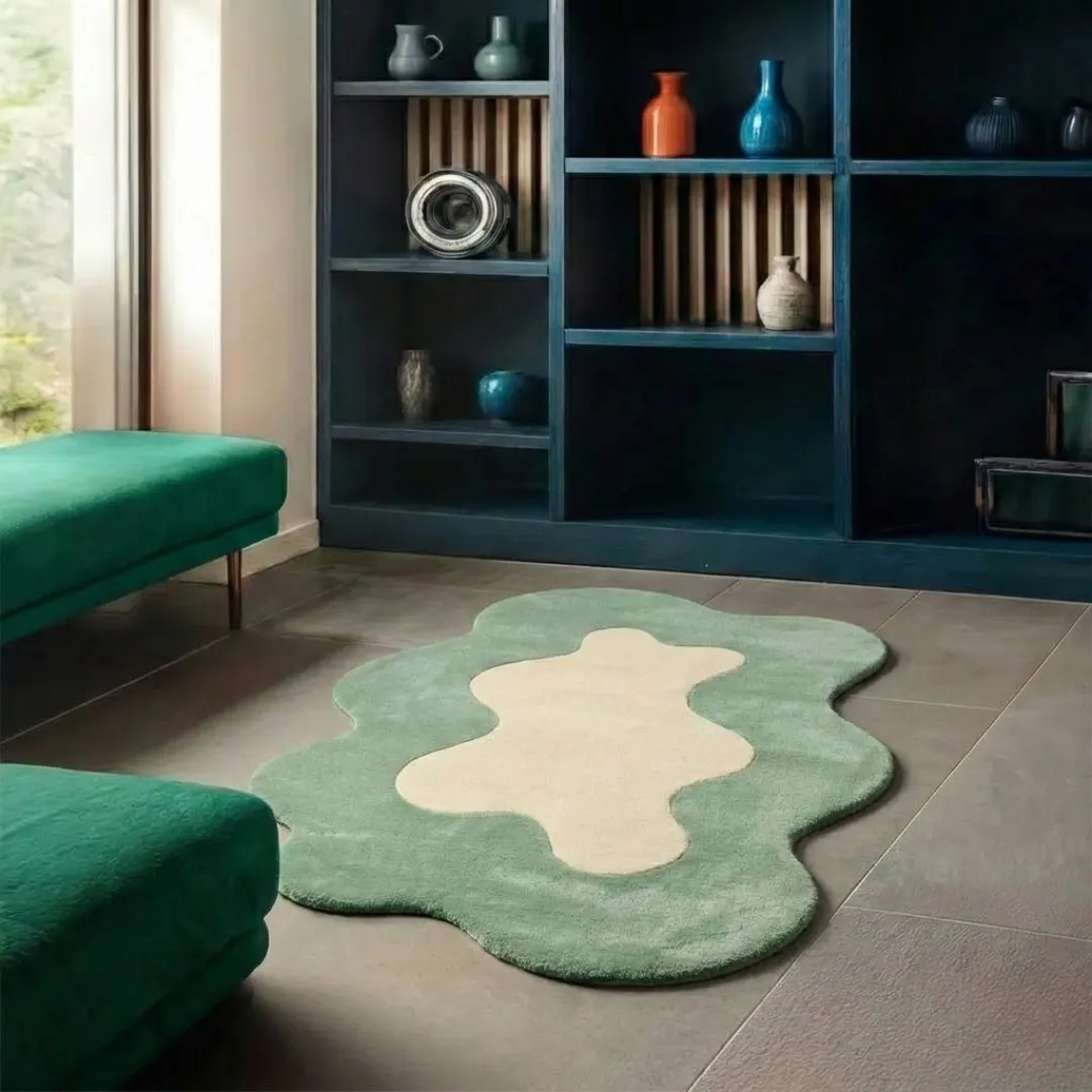 Perspective lifestyle view of the Liom Mist rug placed on a grey tile floor, showcasing its unique wavy border and calming sage green and cream palette in a modern room styled with green velvet benches.