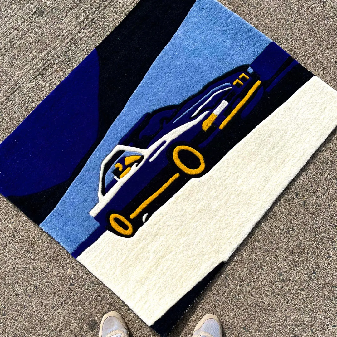 First-person angled lifestyle view of the Noctra Veil rug styled on a concrete street setting, showcasing the vibrant automotive design alongside a pair of sneakers at the bottom edge.