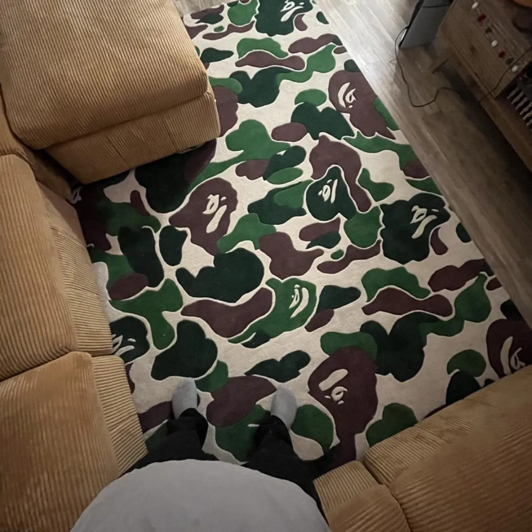 First-person point of view standing on the Shadowfield Camo Terrain rug, styled in a cozy living room next to a tan corduroy sectional sofa, emphasizing the scale of the bold camo pattern.