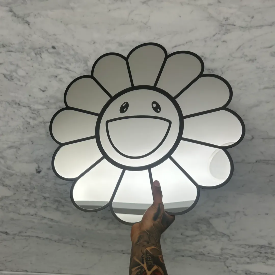 A hand holding up a daisy-shaped mirror with a bold black outlined smiley face in the center, set against a bright white marble background.