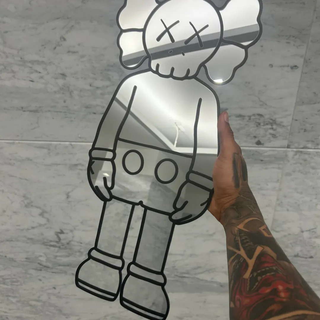 A tattooed hand holding up the full-body Street Bear mirror against a white marble wall, capturing clear reflections of the surrounding room on its glass surface.