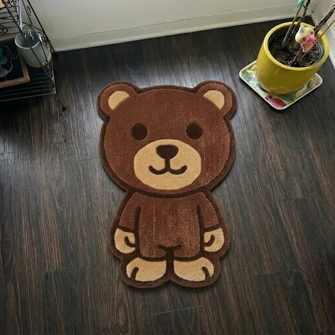 A top-down studio product photograph of a unique, character-shaped area rug featuring a cute brown teddy bear design, displayed on a dark wooden floor.