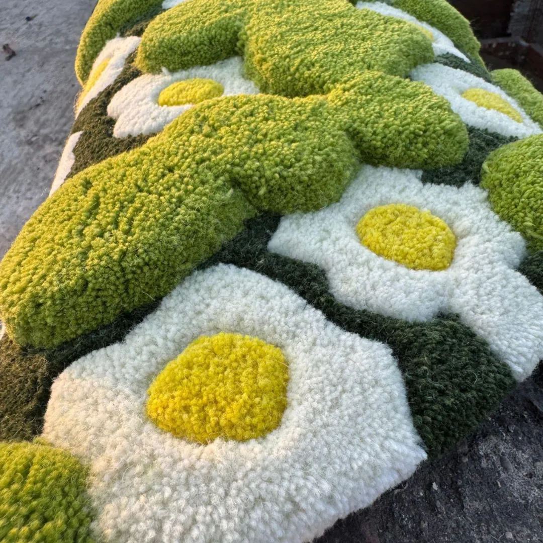 An extreme close-up of the 3-D Moss Rug taken in warm sunlight, highlighting the different pile heights of the tufted yarn. The image focuses on the highly textured, raised bright green moss patches and the plush white daisies with yellow centers.