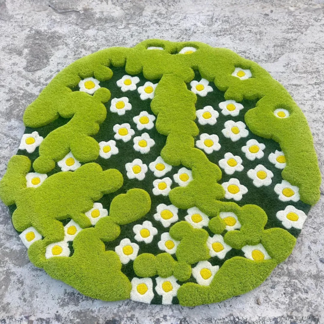 Top-down view of the circular 3-D Moss Rug resting on a light grey concrete surface. The hand-tufted design features raised, bright green mossy shapes overlapping a dark green background filled with white and yellow daisy flowers.