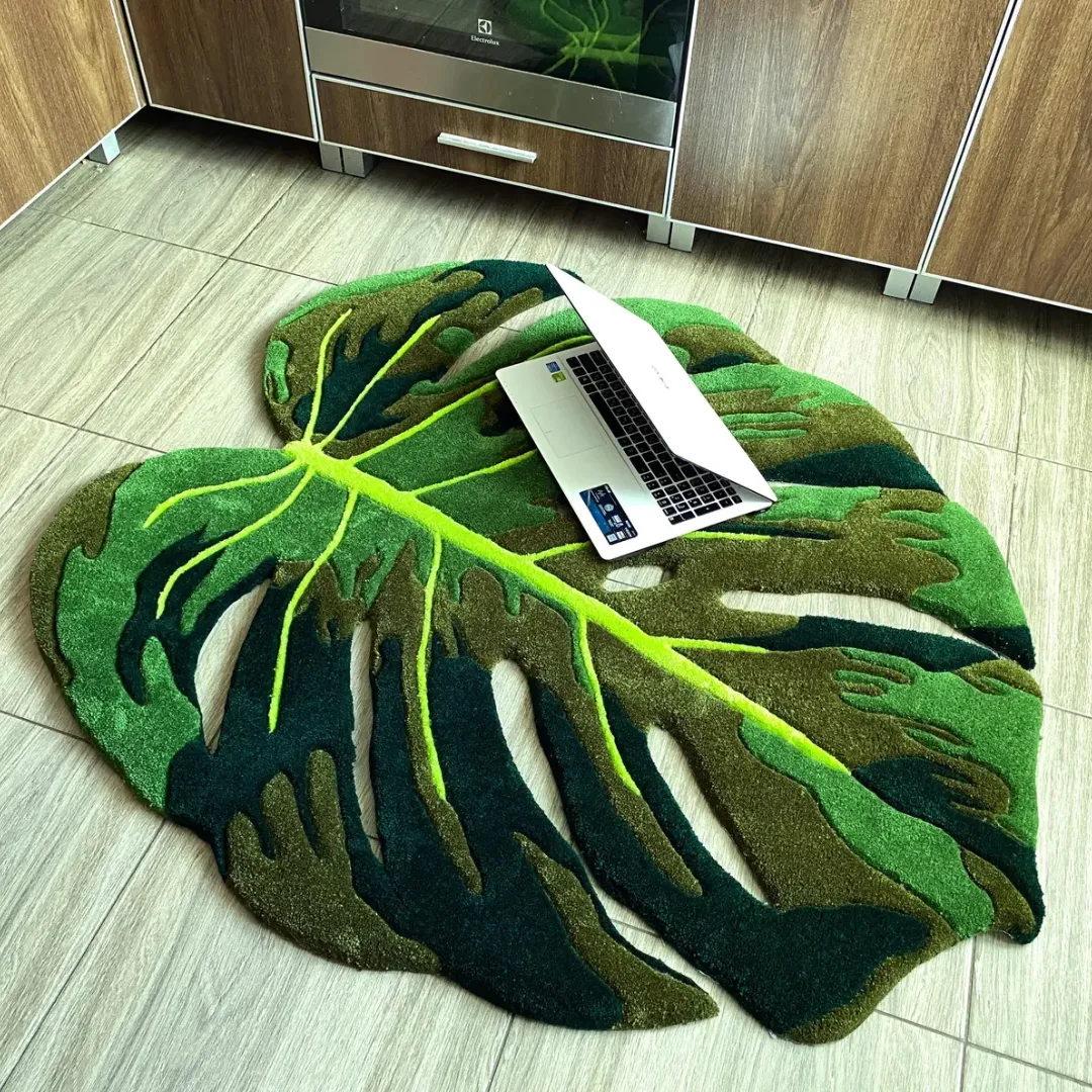 Top-down view of the tufted Monstera Flux Rug on a light tiled floor with a silver laptop resting on it for scale. The rug features a custom cut-out tropical leaf design in multiple shades of green.